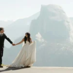 A bride and groom hold hands on a mountain road with beautiful rocky cliffs behind them. The bride wears a long white dress, and the groom wears a black suit. This image connects to the A2 ESL lesson about weddings by showing a different type of ceremony — an outdoor wedding in nature. It helps students talk about where weddings can take place, what people wear, and how weddings can look different in each culture.