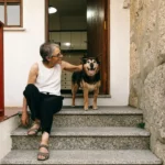 An elderly woman sits on her front steps smiling beside her dog. The image represents the What Does Aging Really Feel Like? B2 ESL lesson from betterclass, where students explore how aging affects the body and mind through vocabulary, video analysis, and reflective discussions about empathy, independence, and life experience.