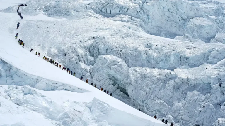 A long line of climbers in colorful gear ascending a steep snow-covered ridge on Mount Everest, with massive glacial ice walls in the background.