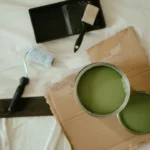 Two open cans of olive green paint with a roller and brush on a drop cloth, ready for a B1 lesson plan on home renovation.