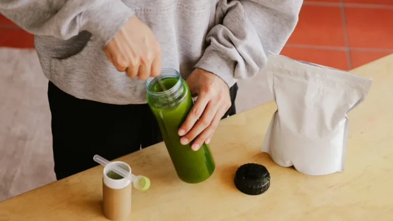 A person stirring a green smoothie in a glass bottle with supplement powder on the table, from an A2 habits lesson.