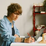 Adult woman taking notes for an A2 lesson plan at a home desk with plants and books nearby.