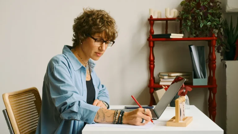 Adult woman taking notes for an A2 lesson plan at a home desk with plants and books nearby.